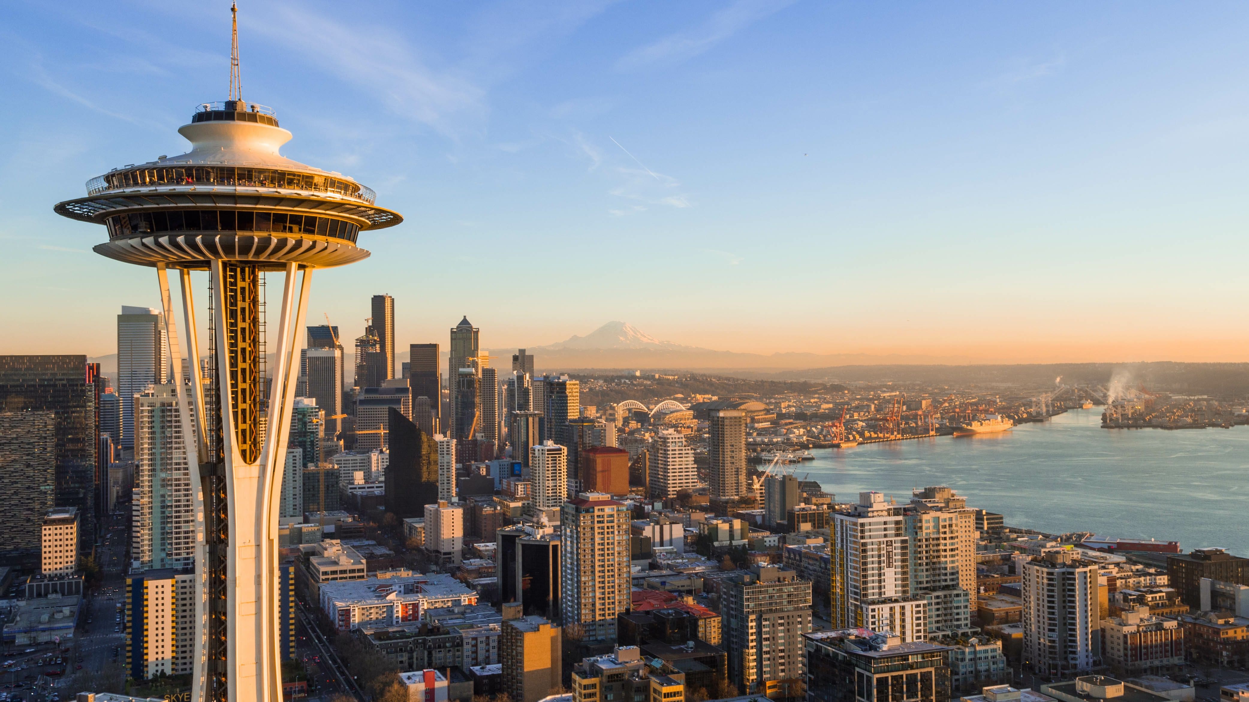 A panoramic view of Seattle’s skyline featuring the Space Needle in the foreground at sunset, with Mount Rainier visible in the distance.