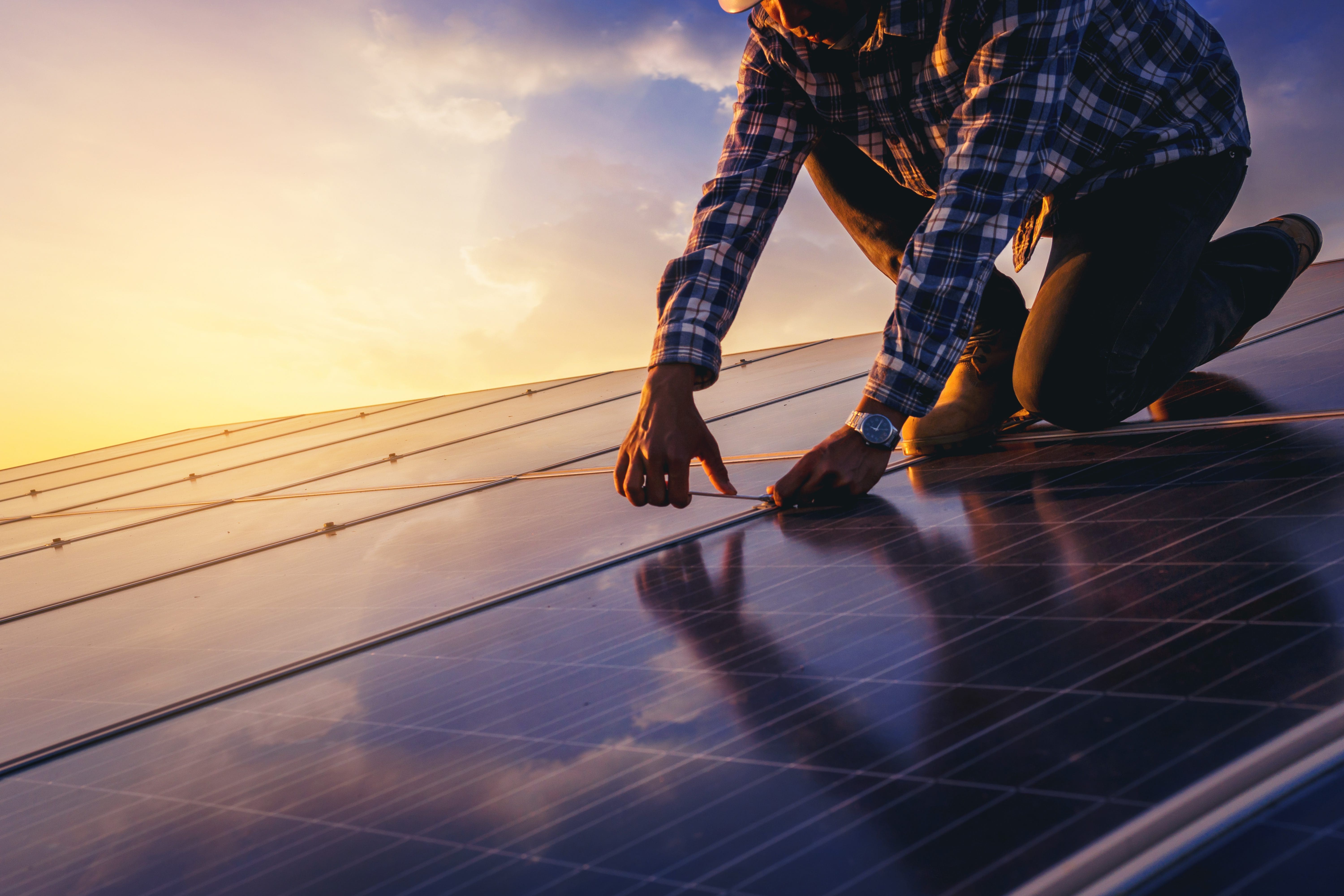 Worker on a rooftop tightening a solar panel module at sunset.